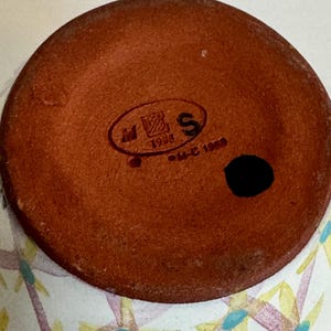 May include: Close-up of the bottom of a ceramic vase. The base is a reddish-brown color with a black spot and a stamped logo. The vase's exterior features a white background with a floral pattern in yellow, purple, and blue.