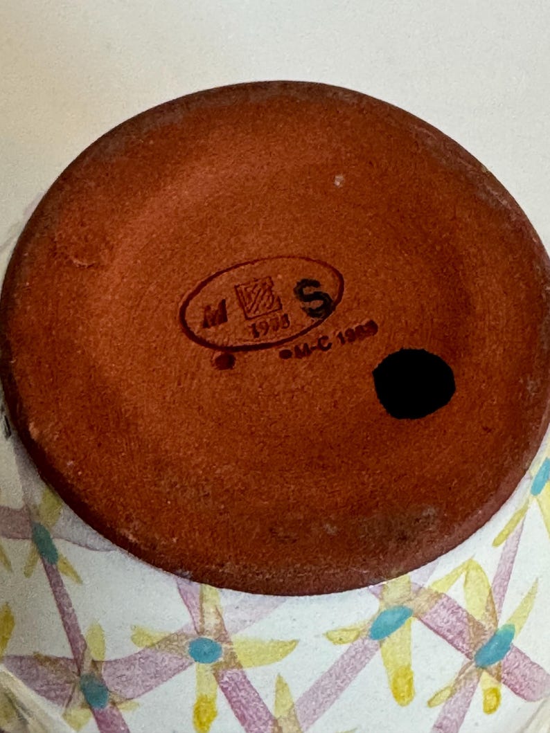 May include: Close-up of the bottom of a ceramic vase. The base is a reddish-brown color with a black spot and a stamped logo. The vase's exterior features a white background with a floral pattern in yellow, purple, and blue.