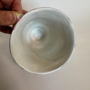 Op de afbeelding: Een handgemaakte keramische mok met een lichtbeige interieur en een licht uitlopende rand. De mok heeft een klein handvat en een gespikkeld exterieur. De binnenkant heeft een gladde, glanzende afwerking.