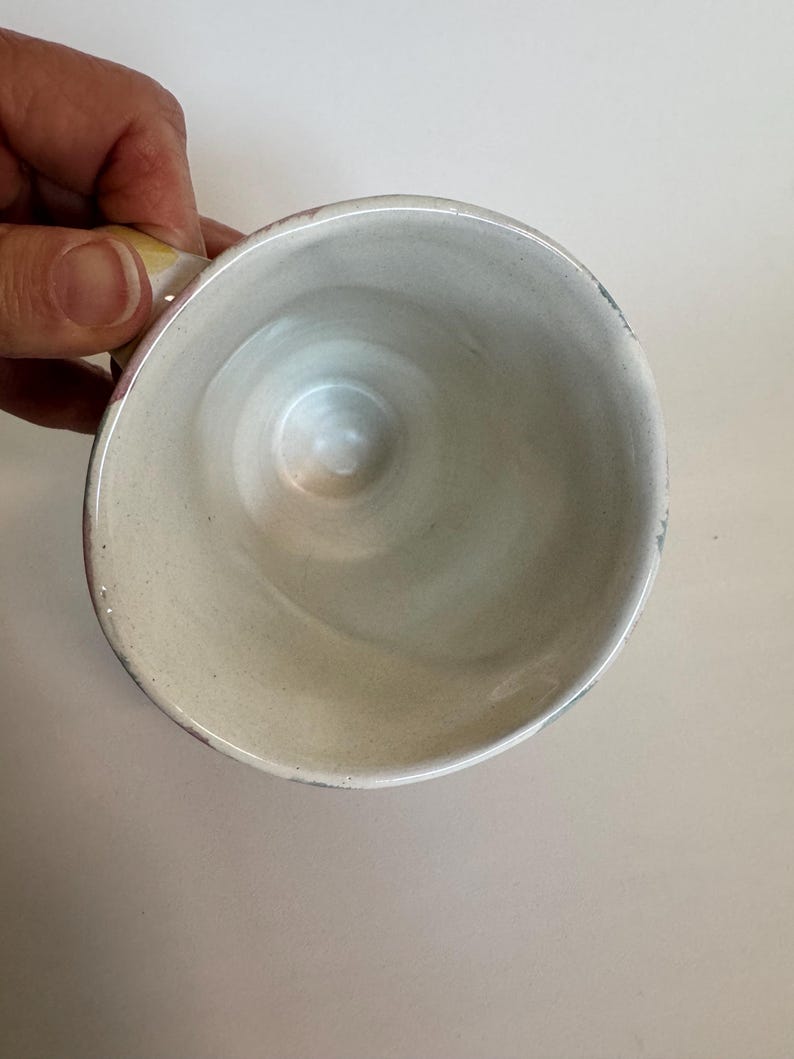 Op de afbeelding: Een handgemaakte keramische mok met een lichtbeige interieur en een licht uitlopende rand. De mok heeft een klein handvat en een gespikkeld exterieur. De binnenkant heeft een gladde, glanzende afwerking.