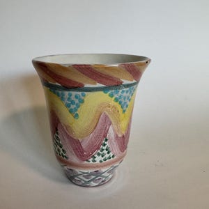 May include: A hand-painted ceramic vase with a flared rim. The vase features a colorful design with yellow, pink, and blue wavy patterns. The base has a geometric design in shades of purple and green. The vase is approximately 6 inches tall.