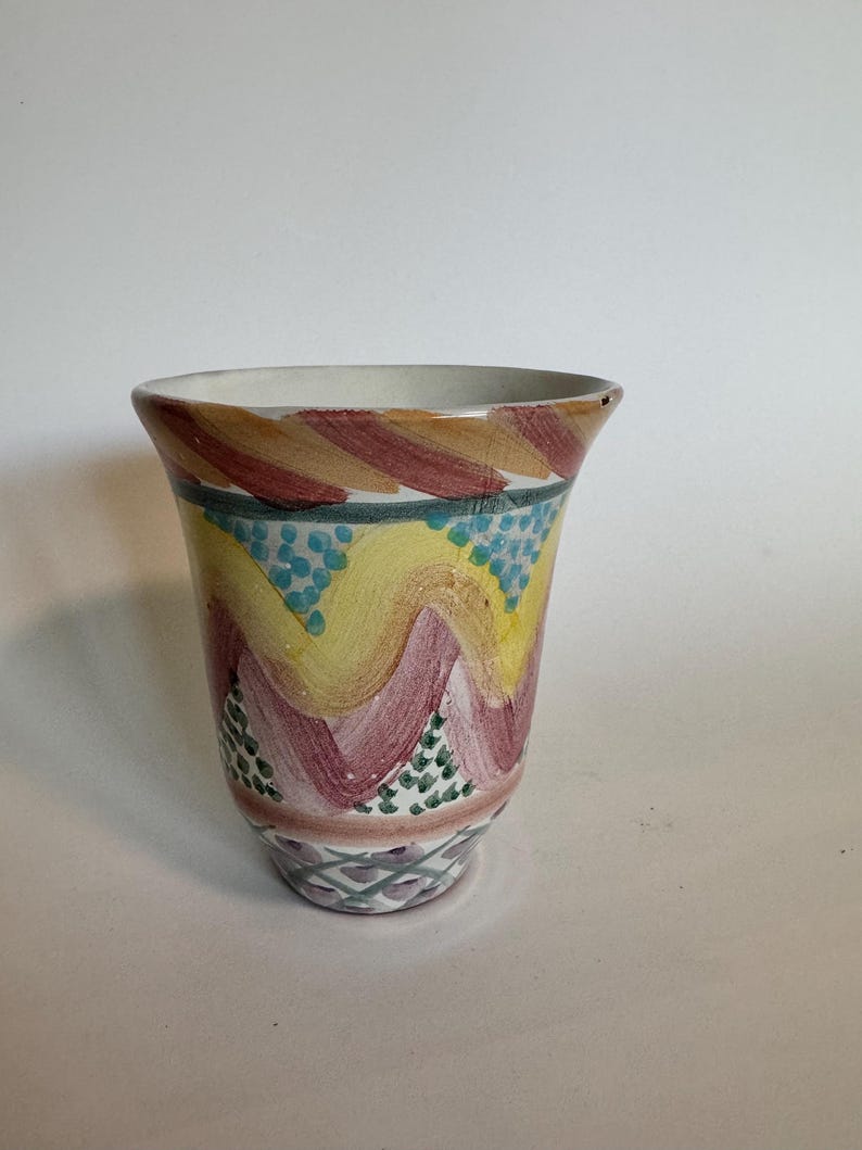 May include: A hand-painted ceramic vase with a flared rim. The vase features a colorful design with yellow, pink, and blue wavy patterns. The base has a geometric design in shades of purple and green. The vase is approximately 6 inches tall.