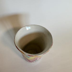 May include: A ceramic vase with a wide, flared opening and a cylindrical body. The interior is a light beige, while the exterior features a colorful, abstract pattern in pink, yellow, and blue. The rim is trimmed with a reddish-brown band.