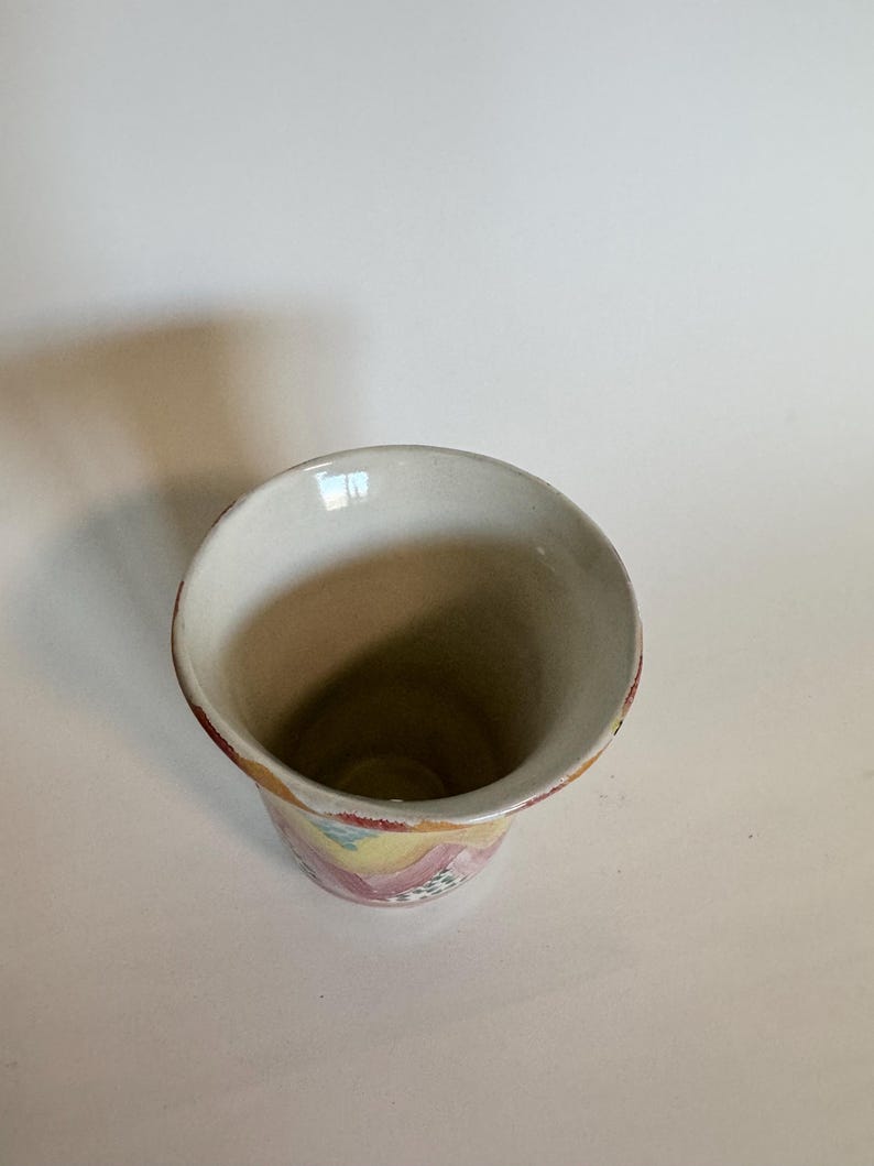 May include: A ceramic vase with a wide, flared opening and a cylindrical body. The interior is a light beige, while the exterior features a colorful, abstract pattern in pink, yellow, and blue. The rim is trimmed with a reddish-brown band.