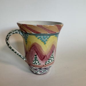 May include: A hand-painted ceramic mug with a flared rim and a curved handle. The mug features a colorful design with yellow, burgundy, and teal zigzags, and green and teal polka dots. The handle is decorated with green dots.
