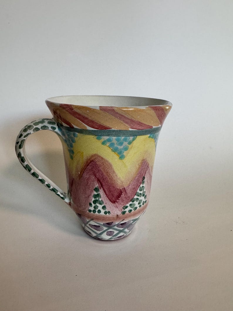 May include: A hand-painted ceramic mug with a flared rim and a curved handle. The mug features a colorful design with yellow, burgundy, and teal zigzags, and green and teal polka dots. The handle is decorated with green dots.
