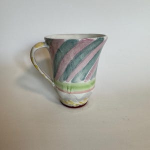 Op de afbeelding: Een keramische mok met een witte binnenkant en een handvat. De buitenkant heeft diagonale strepen in groen, roze en wit. De basis heeft een geel en rood ontwerp. De mok is een uniek stuk drinkgerei.