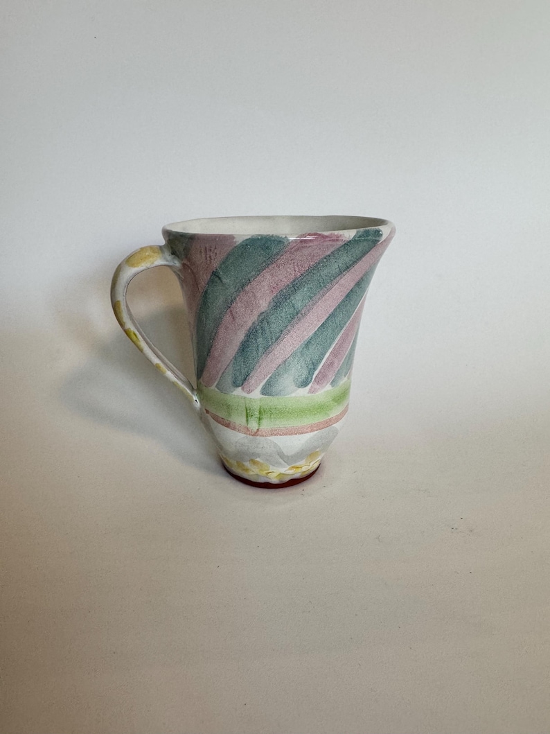 Op de afbeelding: Een keramische mok met een witte binnenkant en een handvat. De buitenkant heeft diagonale strepen in groen, roze en wit. De basis heeft een geel en rood ontwerp. De mok is een uniek stuk drinkgerei.