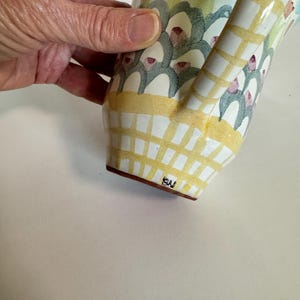 Op de afbeelding: Een handbeschilderde keramische kan met een gebogen handvat. De kan heeft een geel en wit geblokt patroon aan de basis, met bloemen en abstracte ontwerpen in groen, blauw en roze. De basis heeft de initialen "SW".