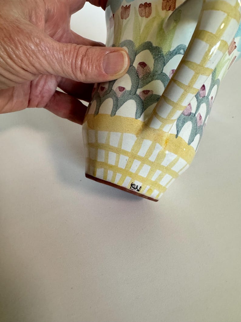 Op de afbeelding: Een handbeschilderde keramische kan met een gebogen handvat. De kan heeft een geel en wit geblokt patroon aan de basis, met bloemen en abstracte ontwerpen in groen, blauw en roze. De basis heeft de initialen "SW".