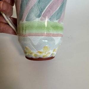 Op de afbeelding: Een keramische mok met een handvat, versierd met diagonale strepen in roze, blauw en groen. De basis heeft een patroon van gele en witte bloemvormen, met een bruine rand.