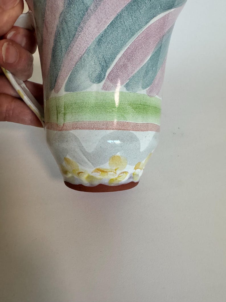 Op de afbeelding: Een keramische mok met een handvat, versierd met diagonale strepen in roze, blauw en groen. De basis heeft een patroon van gele en witte bloemvormen, met een bruine rand.