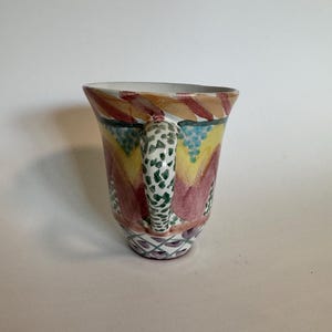 May include: A hand-painted ceramic mug with a colorful design. The mug features a white handle with green spots, and the body is decorated with red, yellow, and blue patterns. The rim is white, and the base has a geometric design.