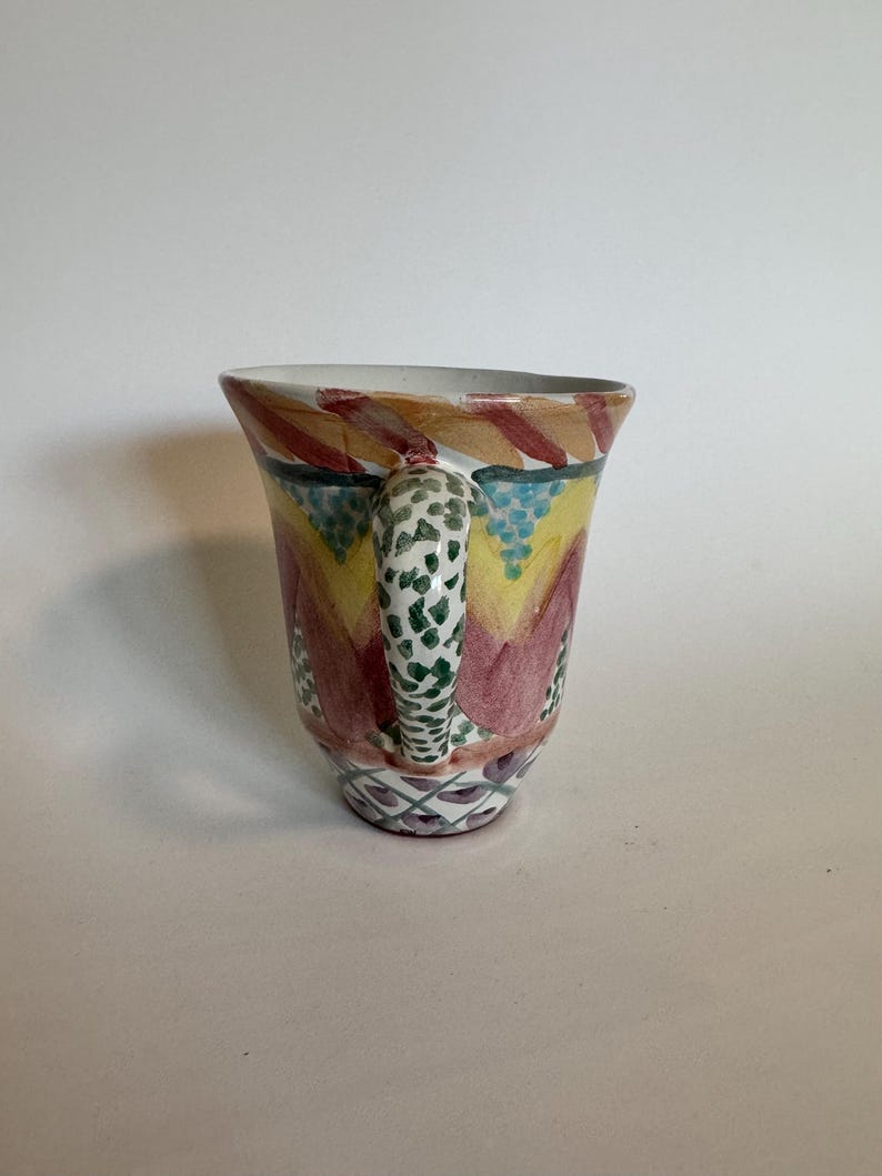 May include: A hand-painted ceramic mug with a colorful design. The mug features a white handle with green spots, and the body is decorated with red, yellow, and blue patterns. The rim is white, and the base has a geometric design.