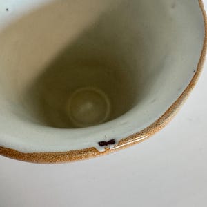 May include: A handmade ceramic mug with a wide opening and a rounded shape. The interior is a light beige color, while the rim has a brown and white speckled design. The mug has a small, dark brown mark on the rim.