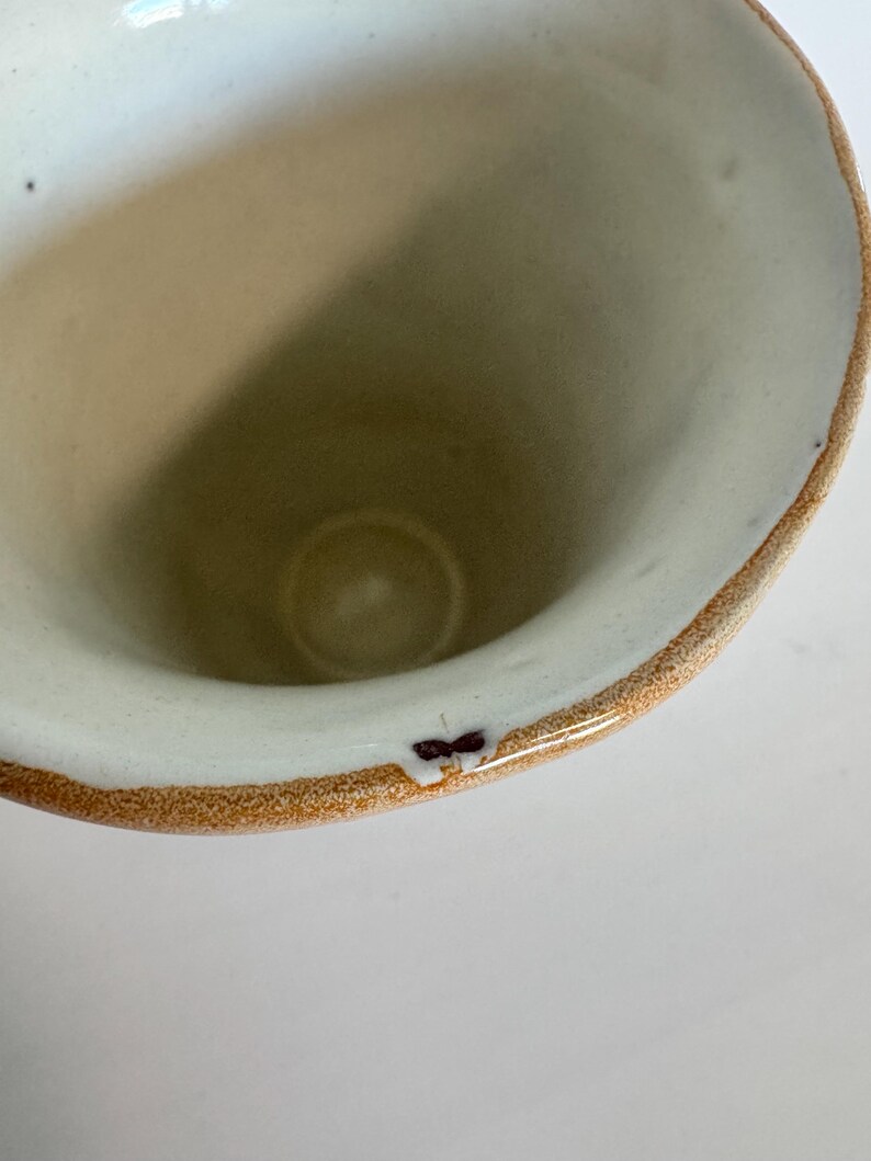 May include: A handmade ceramic mug with a wide opening and a rounded shape. The interior is a light beige color, while the rim has a brown and white speckled design. The mug has a small, dark brown mark on the rim.