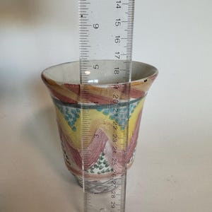 May include: A hand-painted ceramic cup with a colorful design. The cup features a wavy pattern in shades of yellow, pink, and blue. The cup is approximately 3 inches (7.6 cm) in diameter and 4 inches (10.2 cm) tall.