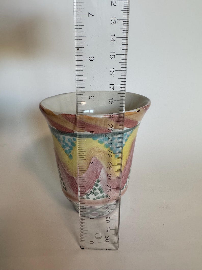 May include: A hand-painted ceramic cup with a colorful design. The cup features a wavy pattern in shades of yellow, pink, and blue. The cup is approximately 3 inches (7.6 cm) in diameter and 4 inches (10.2 cm) tall.