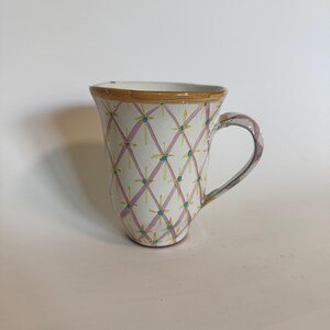 May include: A white ceramic mug with a hand-painted design. The pattern features a grid of pink lines with yellow and blue accents. The rim is trimmed with a brown band, and the handle has a purple and white design.