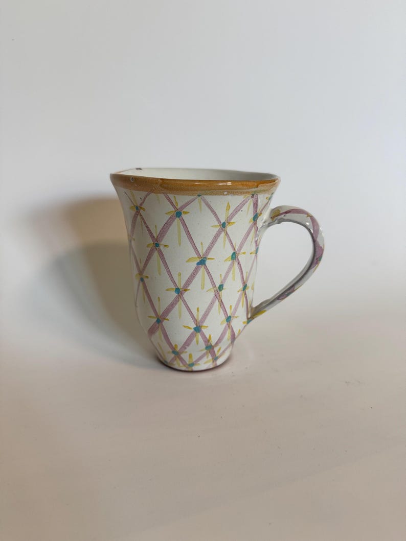 May include: A white ceramic mug with a hand-painted design. The pattern features a grid of pink lines with yellow and blue accents. The rim is trimmed with a brown band, and the handle has a purple and white design.