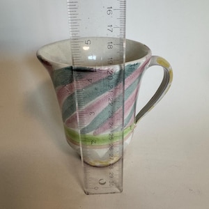 Op de afbeelding: Een kleine, handbeschilderde keramische mok met een oor. De mok heeft een witte binnenkant en een kleurrijke buitenkant met diagonale strepen in roze, groen en geel. De mok is ongeveer 7,6 cm hoog.