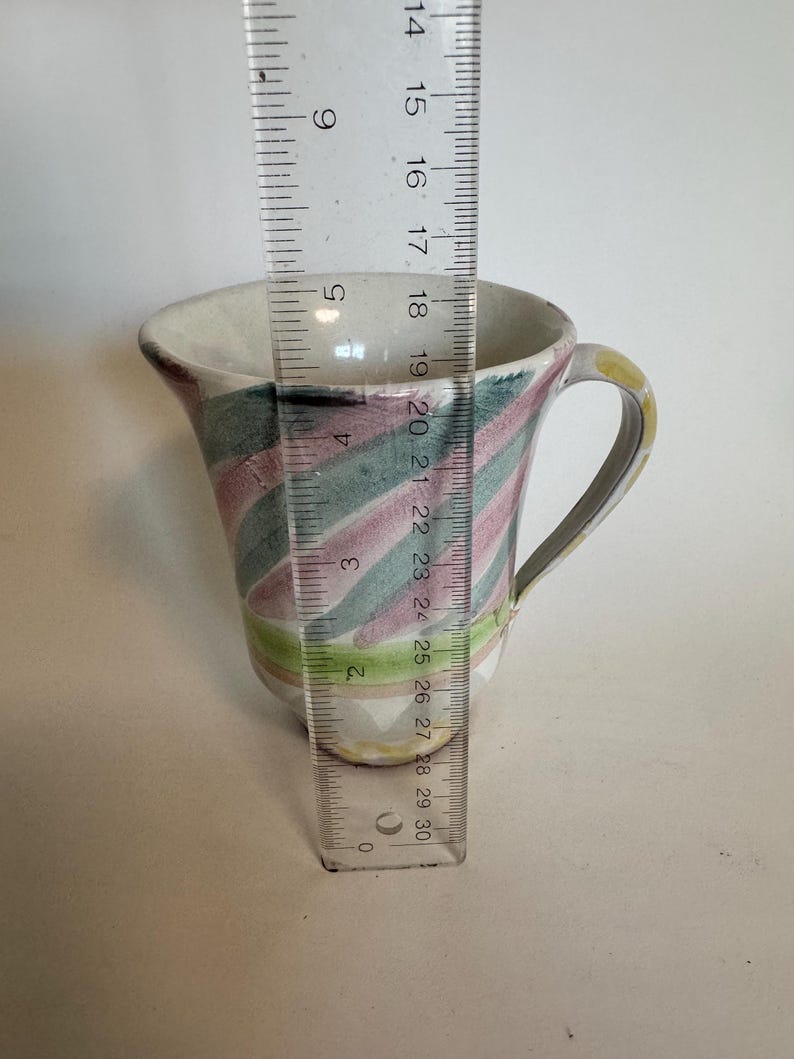 Op de afbeelding: Een kleine, handbeschilderde keramische mok met een oor. De mok heeft een witte binnenkant en een kleurrijke buitenkant met diagonale strepen in roze, groen en geel. De mok is ongeveer 7,6 cm hoog.