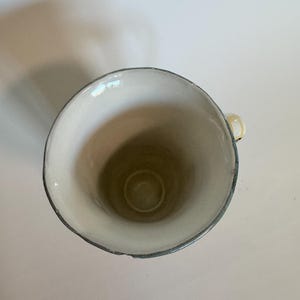 Op de afbeelding: Een witte keramische mok met een grijze rand en een klein handvat. De mok heeft een eenvoudig, elegant ontwerp, perfect om van koffie of thee te genieten. De binnenkant van de mok is lichtbeige.