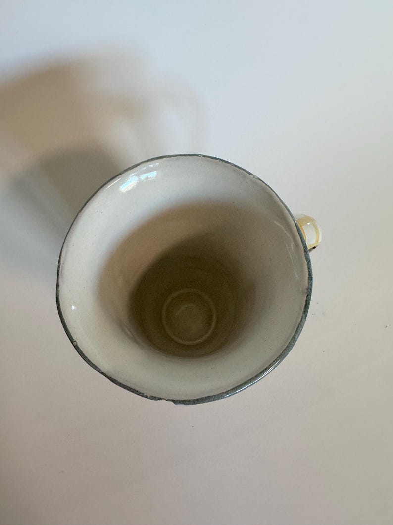Op de afbeelding: Een witte keramische mok met een grijze rand en een klein handvat. De mok heeft een eenvoudig, elegant ontwerp, perfect om van koffie of thee te genieten. De binnenkant van de mok is lichtbeige.