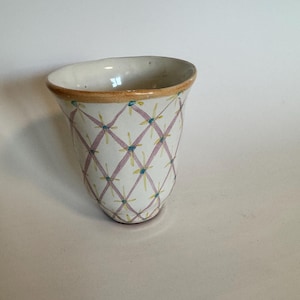 May include: A handmade ceramic vase with a unique design. The vase features a white base adorned with a pink crisscross pattern, accented by yellow and blue floral details. The rim has a brown trim, adding a touch of warmth to the design.