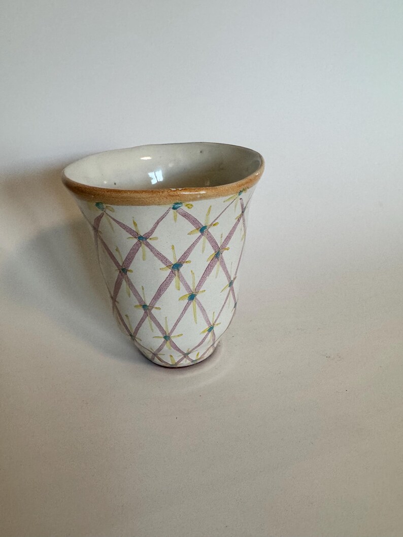 May include: A handmade ceramic vase with a unique design. The vase features a white base adorned with a pink crisscross pattern, accented by yellow and blue floral details. The rim has a brown trim, adding a touch of warmth to the design.