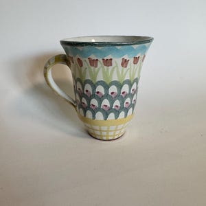 Op de afbeelding: Een keramische mok met een kleurrijk bloemmotief. De mok heeft een blauwe rand, een gele geruite basis en een handvat met gele strepen. De body van de mok is versierd met een patroon van tulpen en bogen in roze, groene en blauwe tinten.