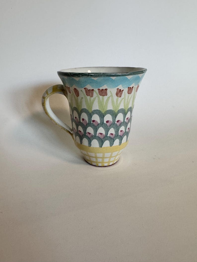 Op de afbeelding: Een keramische mok met een kleurrijk bloemmotief. De mok heeft een blauwe rand, een gele geruite basis en een handvat met gele strepen. De body van de mok is versierd met een patroon van tulpen en bogen in roze, groene en blauwe tinten.