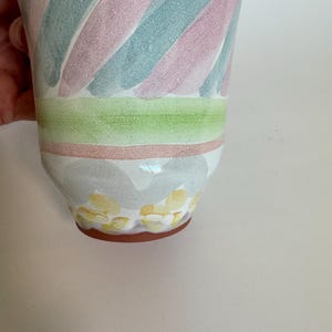 Op de afbeelding: Een keramische vaas met een uitlopende bovenkant, met diagonale strepen in pastelroze, blauwgroen en wit. De vaas heeft een groene en roze band in het midden, met gele bloemaccenten aan de basis.