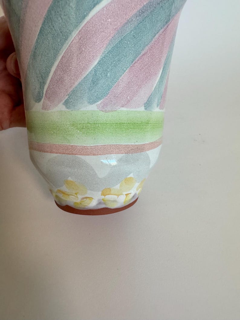 Op de afbeelding: Een keramische vaas met een uitlopende bovenkant, met diagonale strepen in pastelroze, blauwgroen en wit. De vaas heeft een groene en roze band in het midden, met gele bloemaccenten aan de basis.