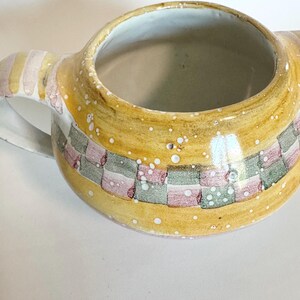 K&ouml;nnte beinhalten: Ein Keramikkrug mit einer weiten &Ouml;ffnung und einem Henkel. Der Krug ist mit einem gelben Hintergrund und einem karierten Muster aus rosa, gr&uuml;nen und grauen Quadraten bemalt. Wei&szlig;e Sprenkel sind &uuml;ber die Oberfl&auml;che verstreut.
