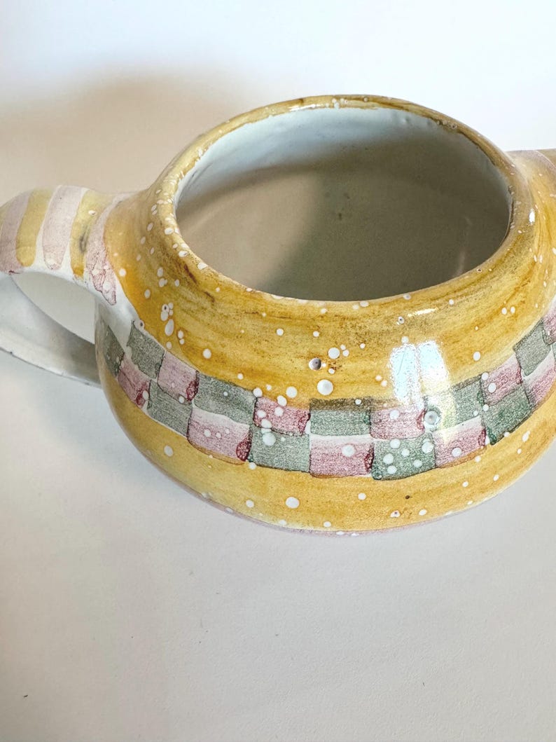 K&ouml;nnte beinhalten: Ein Keramikkrug mit einer weiten &Ouml;ffnung und einem Henkel. Der Krug ist mit einem gelben Hintergrund und einem karierten Muster aus rosa, gr&uuml;nen und grauen Quadraten bemalt. Wei&szlig;e Sprenkel sind &uuml;ber die Oberfl&auml;che verstreut.