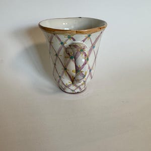 May include: A white ceramic mug with a handle, featuring a lattice pattern in purple and teal, accented with small yellow flowers. The rim is trimmed with a gold-colored band. The mug is designed with a curved shape.