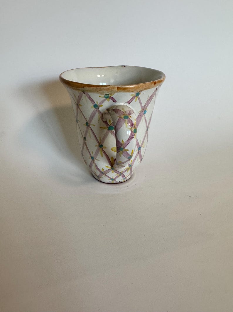 May include: A white ceramic mug with a handle, featuring a lattice pattern in purple and teal, accented with small yellow flowers. The rim is trimmed with a gold-colored band. The mug is designed with a curved shape.