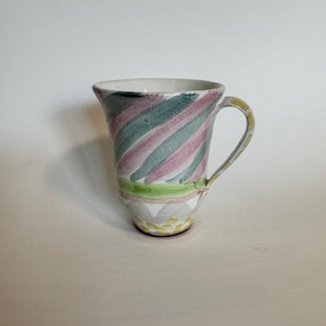 Op de afbeelding: Een keramische kan met een gebogen handvat. De kan heeft een ontwerp van diagonale strepen in groen, roze en wit. De basis heeft een decoratief patroon van geel en grijs. De kan is een decoratief item.