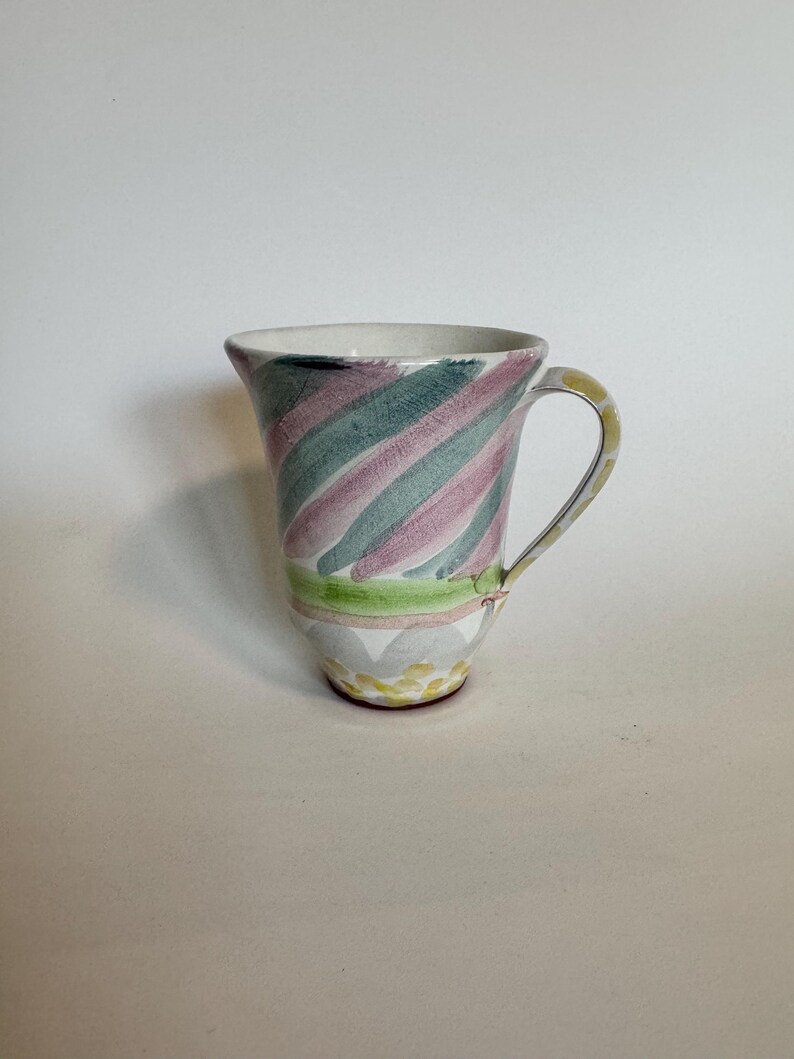 Op de afbeelding: Een keramische kan met een gebogen handvat. De kan heeft een ontwerp van diagonale strepen in groen, roze en wit. De basis heeft een decoratief patroon van geel en grijs. De kan is een decoratief item.