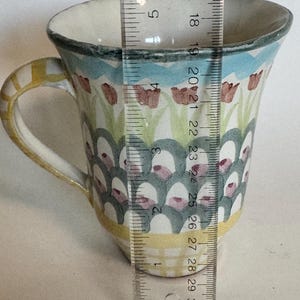 Op de afbeelding: Een keramische mok met een kleurrijk bloemmotief. De mok is ongeveer 10 cm hoog en heeft een gele handgreep en een rand met een blauw en wit patroon. De body van de mok is versierd met rode bloemen en groene bladeren.