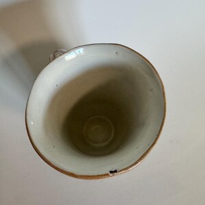 May include: A handmade ceramic mug with a cream-colored interior and a brown rim. The mug has a small handle and a slightly irregular shape, suggesting it is handcrafted. The mug is empty and sits on a white surface.