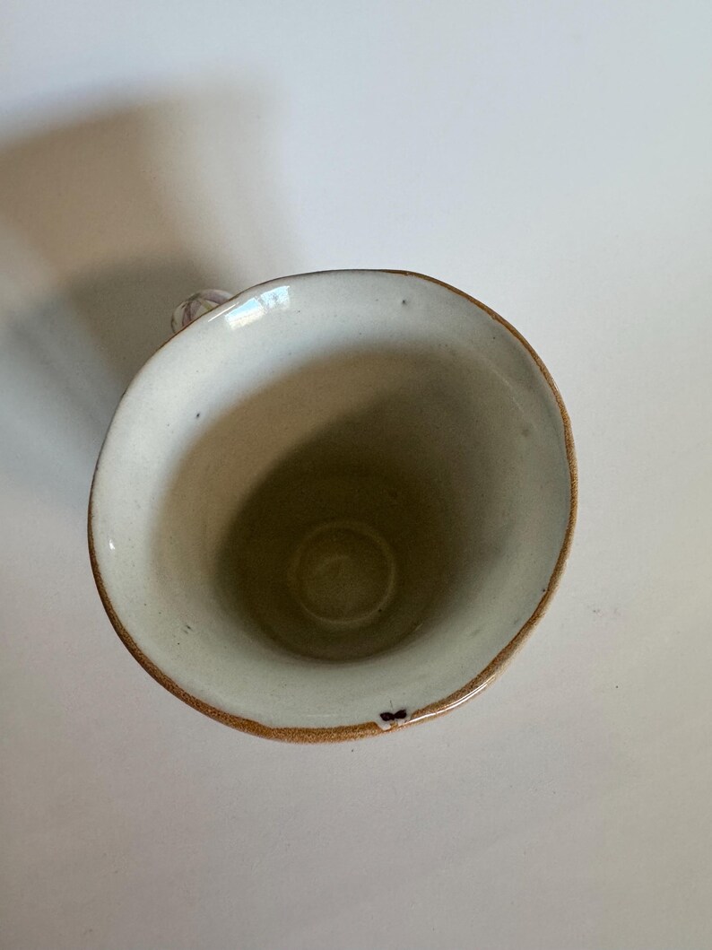 May include: A handmade ceramic mug with a cream-colored interior and a brown rim. The mug has a small handle and a slightly irregular shape, suggesting it is handcrafted. The mug is empty and sits on a white surface.