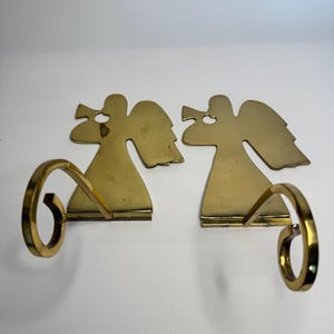 May include: A pair of polished brass wall sconces featuring a flat, silhouette-style angel figure playing a trumpet. Each sconce includes a curved, circular arm extending forward from the base of the angel, designed to hold a candle or small item.