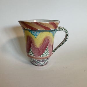 May include: A hand-painted ceramic mug with a flared rim. The mug features a colorful design with red, yellow, and green accents. The handle is speckled green, and the base has a geometric pattern. A unique piece for coffee or tea.