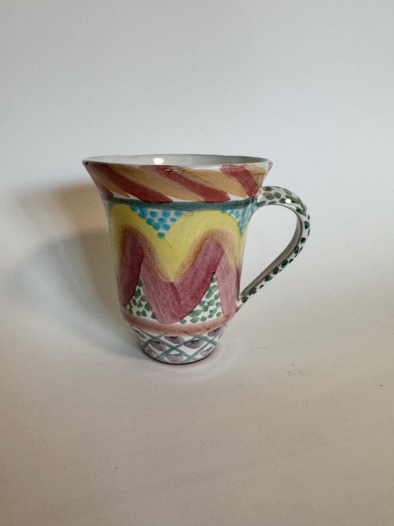 May include: A hand-painted ceramic mug with a flared rim. The mug features a colorful design with red, yellow, and green accents. The handle is speckled green, and the base has a geometric pattern. A unique piece for coffee or tea.