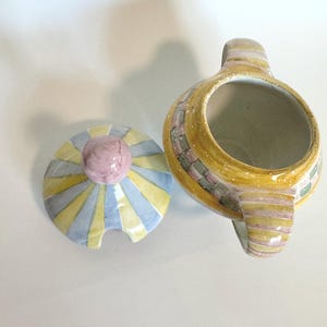 K&ouml;nnte beinhalten: Eine Zuckerdose aus Keramik mit passendem Deckel. Die Sch&uuml;ssel ist gelb mit einem Mosaikmuster, und der Deckel hat ein Sonnenstrahl-Design in Gelb, Blau und Grau. Der Deckel hat einen rosa Knauf. Die Zuckerdose hat einen Griff.