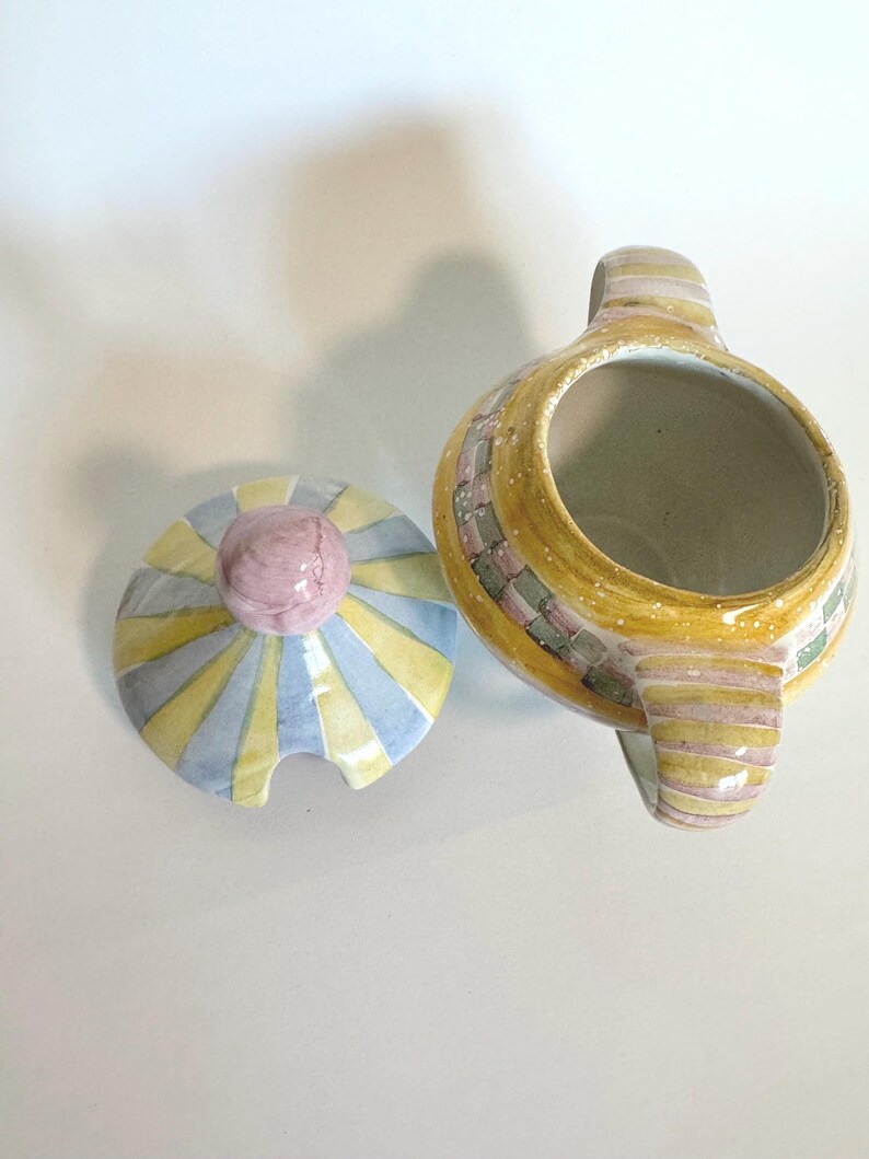 K&ouml;nnte beinhalten: Eine Zuckerdose aus Keramik mit passendem Deckel. Die Sch&uuml;ssel ist gelb mit einem Mosaikmuster, und der Deckel hat ein Sonnenstrahl-Design in Gelb, Blau und Grau. Der Deckel hat einen rosa Knauf. Die Zuckerdose hat einen Griff.