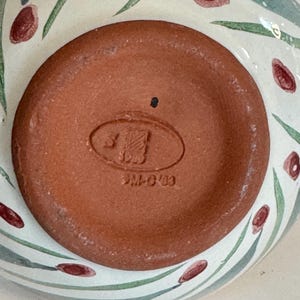 K&ouml;nnte beinhalten: Nahaufnahme des Bodens einer Keramikschale. Der Schalenboden ist rotbraun mit einem gepr&auml;gten Stempel. Die Au&szlig;enseite der Schale hat einen wei&szlig;en Hintergrund mit gr&uuml;n und rot bemalten floralen Designs.