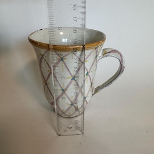 May include: A white ceramic mug with a decorative pattern of intersecting lines and small blue and pink dots. The rim is trimmed with a gold-colored band. The mug is approximately 4 inches tall.